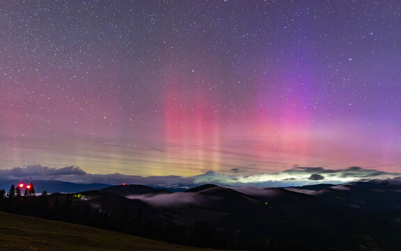 View of the aurora borealis illuminating the night sky above the Graz Highlands with low-lying fog and dark mountain silhouettes in Bruck-Murzzuschlag District, Styria, Austria.