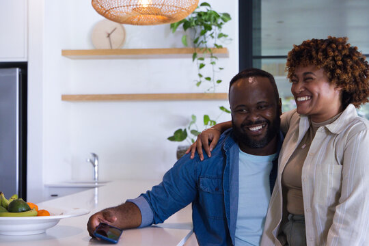 African American couple leaning on white island in kitchen holding smartphone, smiling and laughing