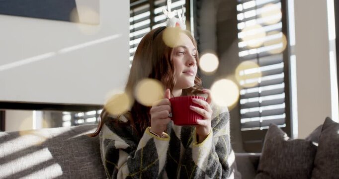 Warm bokeh drifting adult female sitting on couch in antler headband holding red mug gazing outside