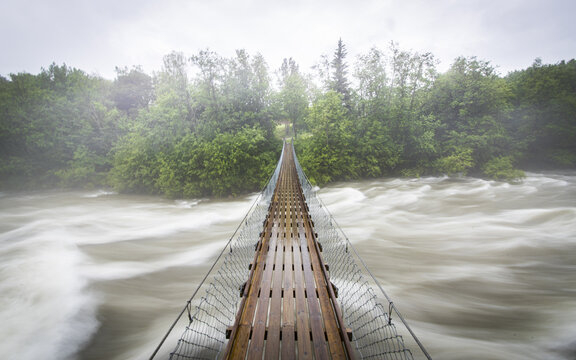 View of a wooden suspension bridge crossing a turbulent, fast-flowing river surrounded by lush green forest in Junkerdal, Nordland, Norway.
