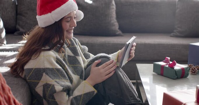 Female sitting on sofa wearing Santa hat tapping tablet replying to holiday messages near gifts