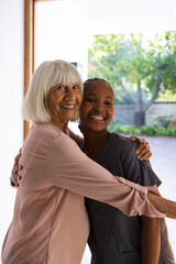 Diverse women hugging at wooden door frame, Senior in light pink blouse holding nurse in scrubs © WavebreakMediaMicro