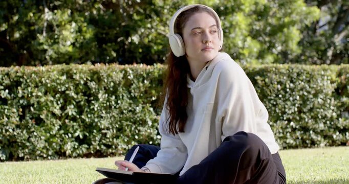 Woman sitting on blanket beside hedge, sketching on tablet with stylus and headphones