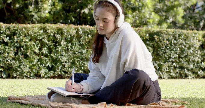 Female in her 20s sitting on blanket in park with headphones writing notes in notebook