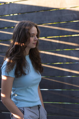 Fototapeta premium Woman wearing blue tee, standing hands in pockets on patio by wood fence, sunlight striping hair