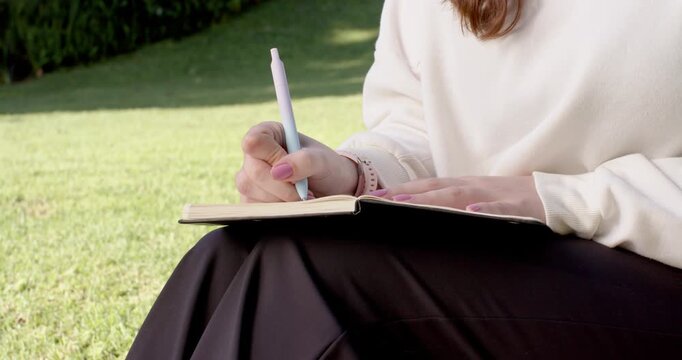 Pen poised, woman is journaling on lap in park with left hand holding notebook page