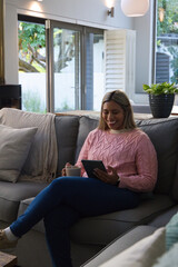 Naklejka premium Mid-adult African American woman sitting on grey sofa in living room holding tablet and mug