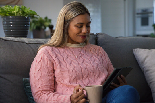 Mid adult African American woman sitting on sofa in living room using tablet and holding mug