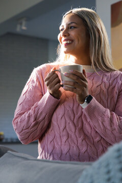 African American woman holding mug, wearing pink sweater in kitchen smiling gazing right