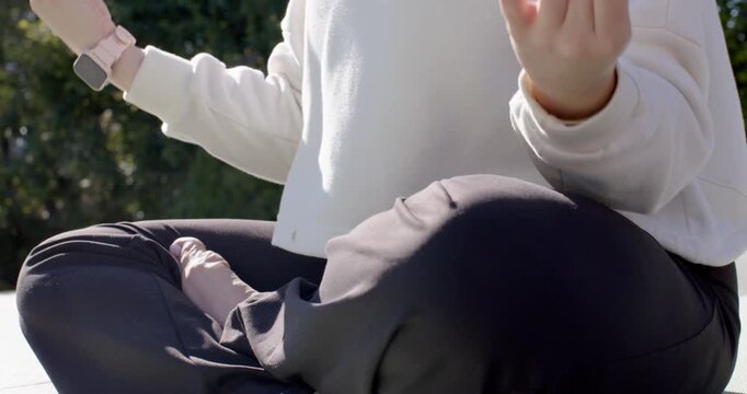 Woman is adjusting hips, settling cross-legged on pale mat near hedge, forming mudras to meditate