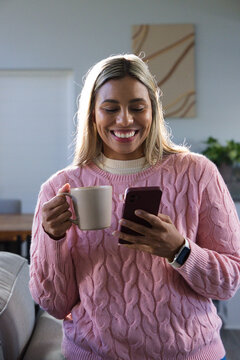 African American woman in pink sweater smiling while holding maroon phone and mug in living room