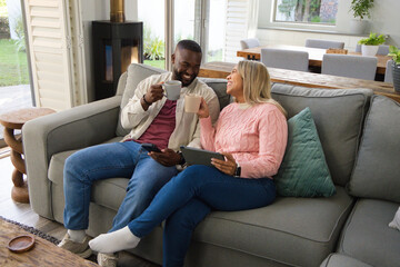 Naklejka premium Diverse couple sitting on gray sofa in living room holding mugs, tablet, smartphone, smiling