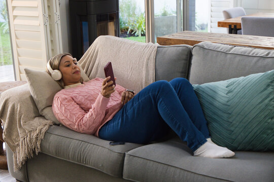 African woman reclining on gray sofa in living room, wearing white headphones, holding maroon phone