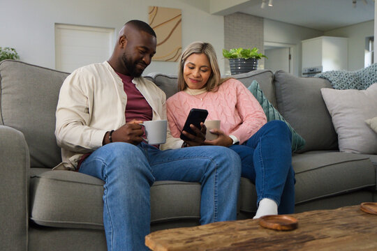 Couple sitting on gray sofa holding ceramic mugs and looking at smartphone in living room
