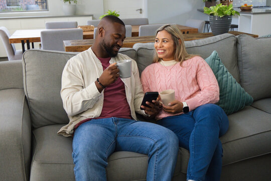 African American couple sitting on grey sofa in modern home holding mugs and smartphone