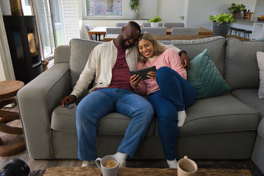 African couple sitting on grey sofa in living room, leaning and viewing tablet with mugs