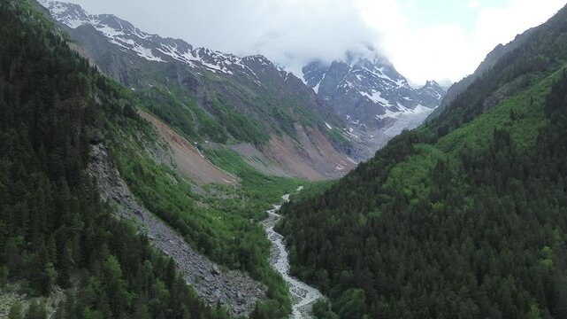 Static 4K drone footage flies backward in Tekhuri gorge, Georgia. Dense green forest lines one side while bare rocky slopes line the other beneath snow-patched, cloud-veiled peaks.