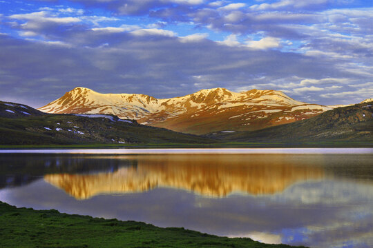 View of Sheosar Lake with snow-capped mountains reflecting in the calm water under a cloudy sky in Deosai National Park, Gilgit Baltistan, Pakistan.