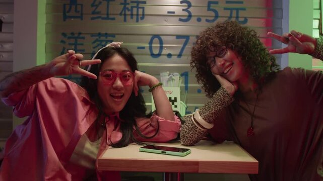 Portrait of Asian young adult woman and Caucasian young adult woman sitting at cafe table, posing with peace signs. Clip is suitable for youth culture and diverse lifestyle marketing campaigns
