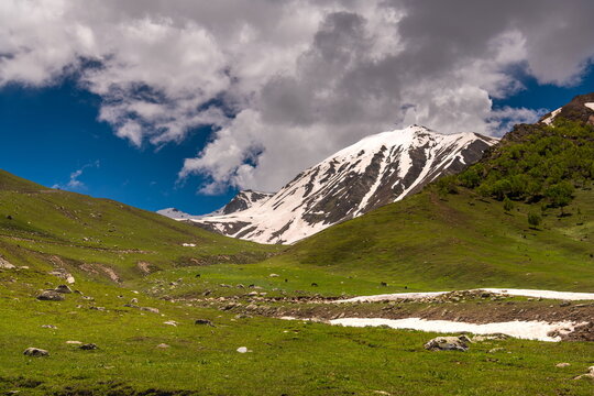View of the lush green meadows and snow-capped peaks of Minimarg Astore, with rolling hills, melting snow, and a dramatic cloudy sky in District Astore, Gilgit Baltistan, Pakistan.