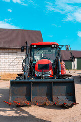 Red farm tractor with front loader isolated in a rural farmyard, modern agricultural machinery parked near barn buildings under a bright blue sky © Iryna
