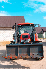 Red farm tractor with front loader isolated in a rural farmyard, modern agricultural machinery parked near barn buildings under a bright blue sky © Iryna
