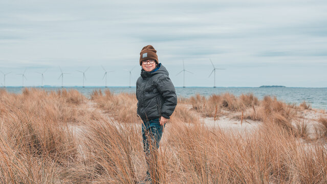 Smiling boy stands in dune grass overlooking offshore turbines.