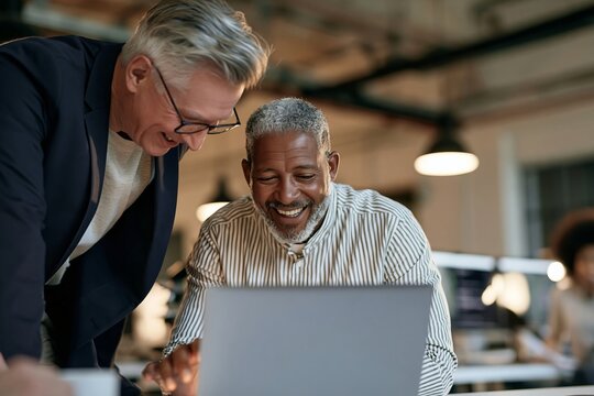 Two mature business colleagues smiling while looking at a laptop screen in a modern office space
