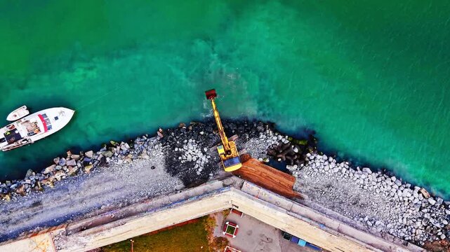 Drone view of dredging excavator and boat. Top down shot of a yellow crane arm reaching into green sea beside an anchored boat.
