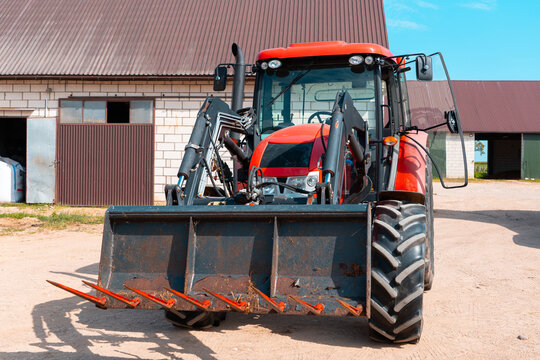 Red farm tractor with front loader isolated in a rural farmyard, modern agricultural machinery parked near barn buildings under a bright blue sky