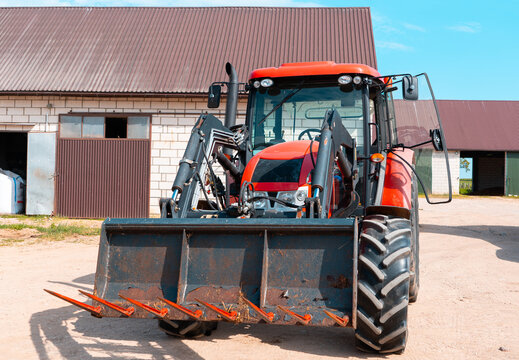 Red farm tractor with front loader isolated in a rural farmyard, modern agricultural machinery parked near barn buildings under a bright blue sky