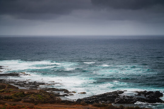 View of the rugged coastline at Mirador Punta Del Hidalgo with crashing waves against dark volcanic rocks under a dramatic cloudy sky in La Laguna, Canarias, Spain.