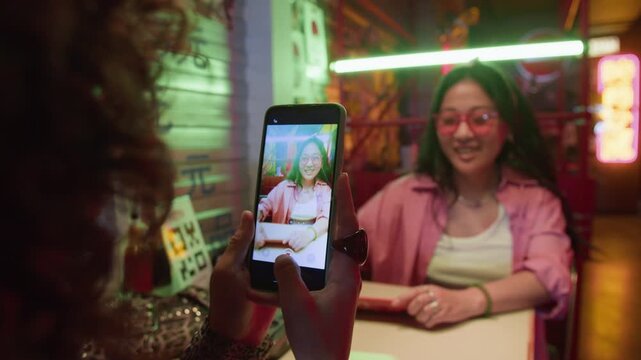 Over shoulder shot of young adult Asian woman posing while friend recording her on smartphone in neon-lit cafe. Footage is suitable for social media marketing and mobile app promotions