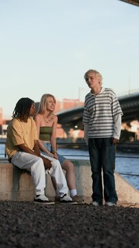 Diverse teenagers, including African American male and Caucasian female, sitting by river and talking with friend. Perfect for youth lifestyle and diversity marketing campaigns, vertical