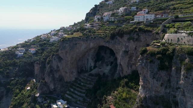 A large cave set on a cliff near houses on the Amalfi Coast. The sun shines on the clear water and the steep land. People can be seen walking along paths.
