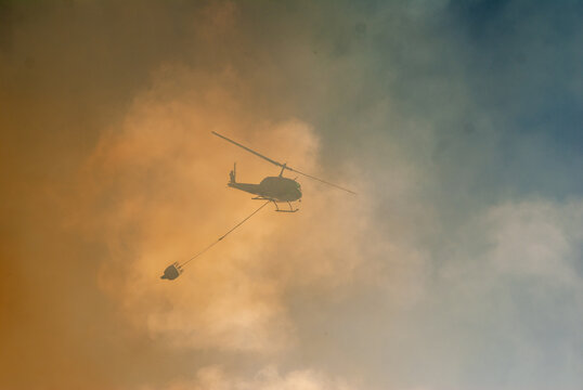 View of a firefighting helicopter carrying a water bucket through thick smoke during a wildfire operation in South Africa.