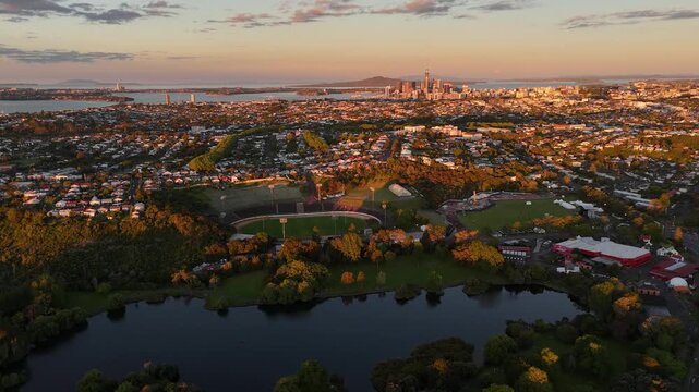 Bird&rsquo;s-eye aerial view over Auckland&rsquo;s Mount Albert suburbs at sunset, revealing residential neighborhoods, green spaces, waterfront, and the city skyline in golden evening light.