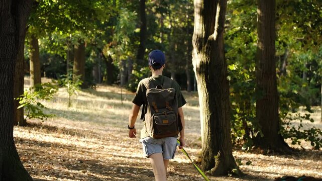 Woman with a backpack walks an Australian Shepherd through a wooded park as the dog sniffs near a tree. Pet care, companionship and active outdoor lifestyle