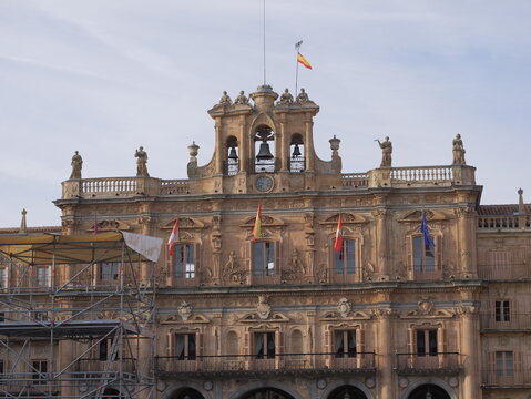 View of the ornate facade of the Plaza Mayor, featuring stone carvings, a clock tower with bells, and Spanish flags, with scaffolding on the left, Salamanca, Castile and Leon, Spain.