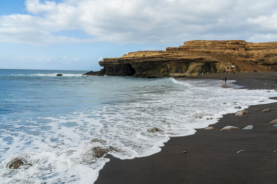 View of the black sand beach and dramatic limestone cliffs of the Ajuy caves, with gentle waves rolling onto the shore in Ajuy, Fuerteventura, Spain.