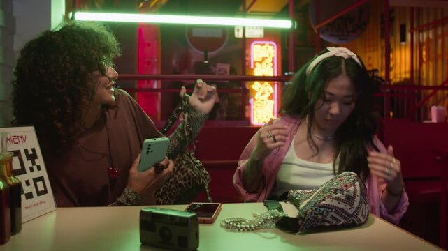 Two young adult Asian women sitting at cafe table, talking and looking at smartphone in neon-lit restaurant. Footage is ideal for youth lifestyle, modern friendship, and mobile app commercials