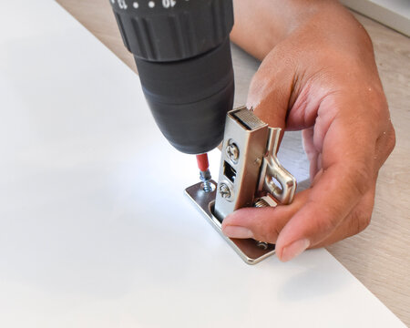 Close-up of a carpenter's hand using an electric drill to screw a metal concealed cabinet hinge onto a white board