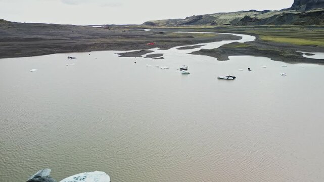 Aerial view across a calm lagoon with small ice fragments and braided river channels in South Iceland, surrounded by dark volcanic plains and distant mountains 4K.