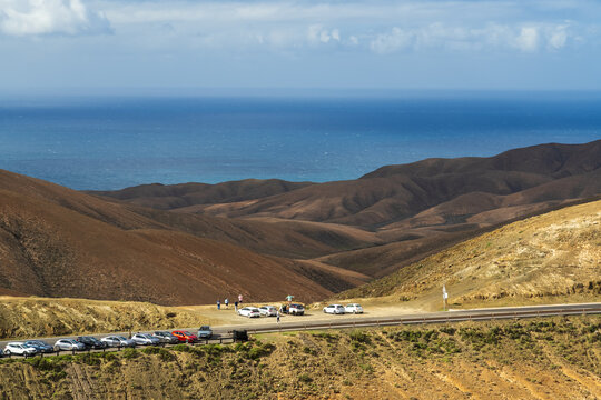 View of a scenic viewpoint with parked cars and people overlooking the rolling arid hills and the blue ocean in Pajara, Canary Islands, Spain.