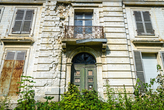 Entr&eacute;e et le premier &eacute;tage d'une demeure bourgeoise en ruine, montrant une ma&ccedil;onnerie en pierre calcaire &eacute;croul&eacute;e, un balcon ouvrag&eacute; rouill&eacute; et une porte envahie par la v&eacute;g&eacute;tation.