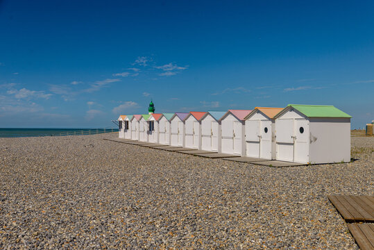 Perspective le long d'une enfilade de cabines de plage blanches aux toitures pastel, avec le phare &agrave; d&ocirc;me vert visible en arri&egrave;re-plan sur une vaste &eacute;tendue de galets sous un ciel azur immacul&eacute;.
