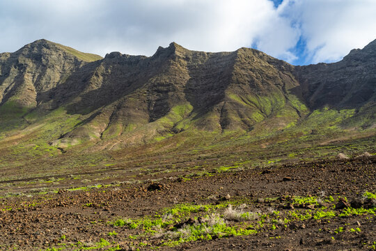 View of the rugged volcanic mountains of Cofete with patches of green vegetation and rocky terrain under a cloudy sky in Cofete, Canary Islands, Spain.