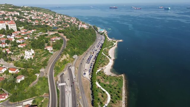 Aerial view of the Koper coastline in Slovenia featuring a coastal road, beach, Adriatic Sea, ships offshore and a green hillside in a scenic urban coastal landscape.