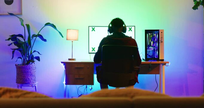 A back-shot view of a young gamer playing games. Online video game on his powerful green screen computer. The room and PC have multi-colored RGB led lights inside the CPU and a lamp on his desk.
