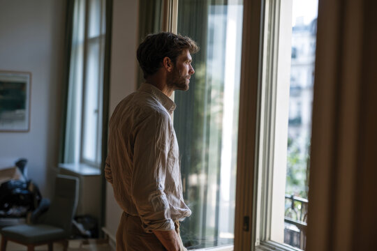 A reflective man stands by the window, bathed in warm sunlight, lost in thought, creating a mood of contemplation, serenity, and connection to the outside world.